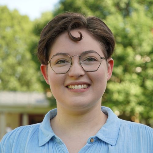 a nonbinary person with glasses and short curly brown hair smiling