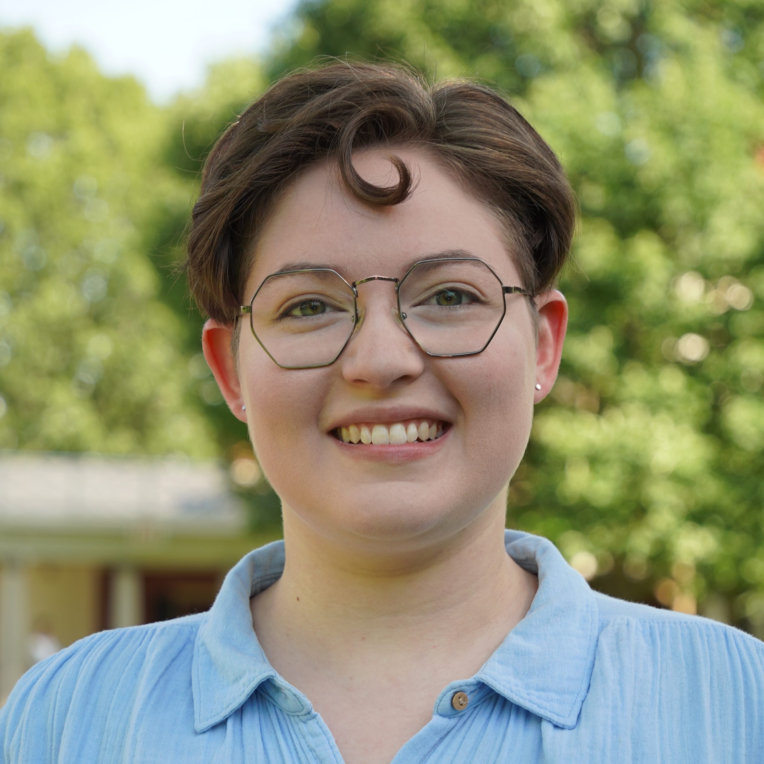 a nonbinary person with glasses and short curly brown hair smiling