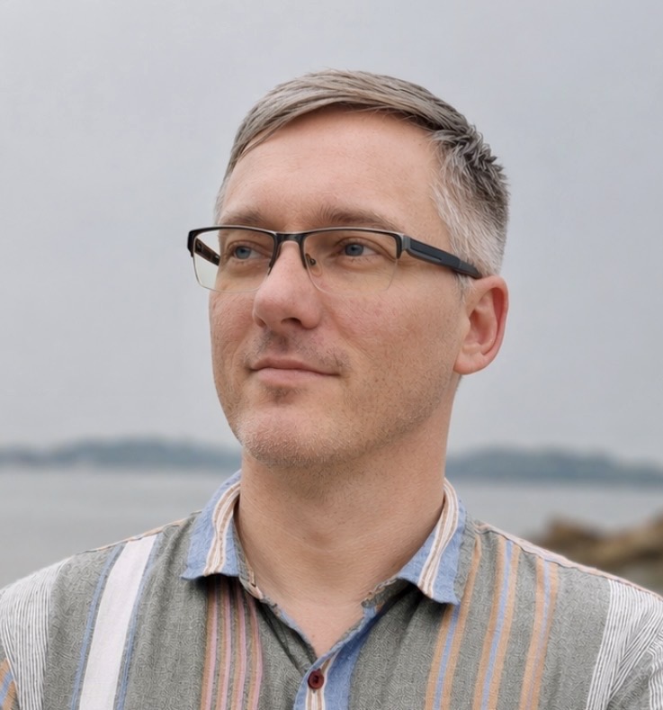 a white man with black-framed glasses looking off to the left in front of a gray sky