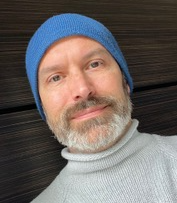 a man with a roan beard, blue beanie, and pale grey sweater stands before a dark wood paneled wall facing directly at the camera in an image that is slightly tilted left