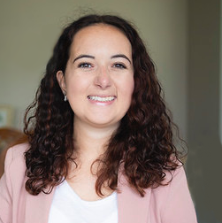 A woman with curly brown hair resting on her shoulders wearing a pink blazer